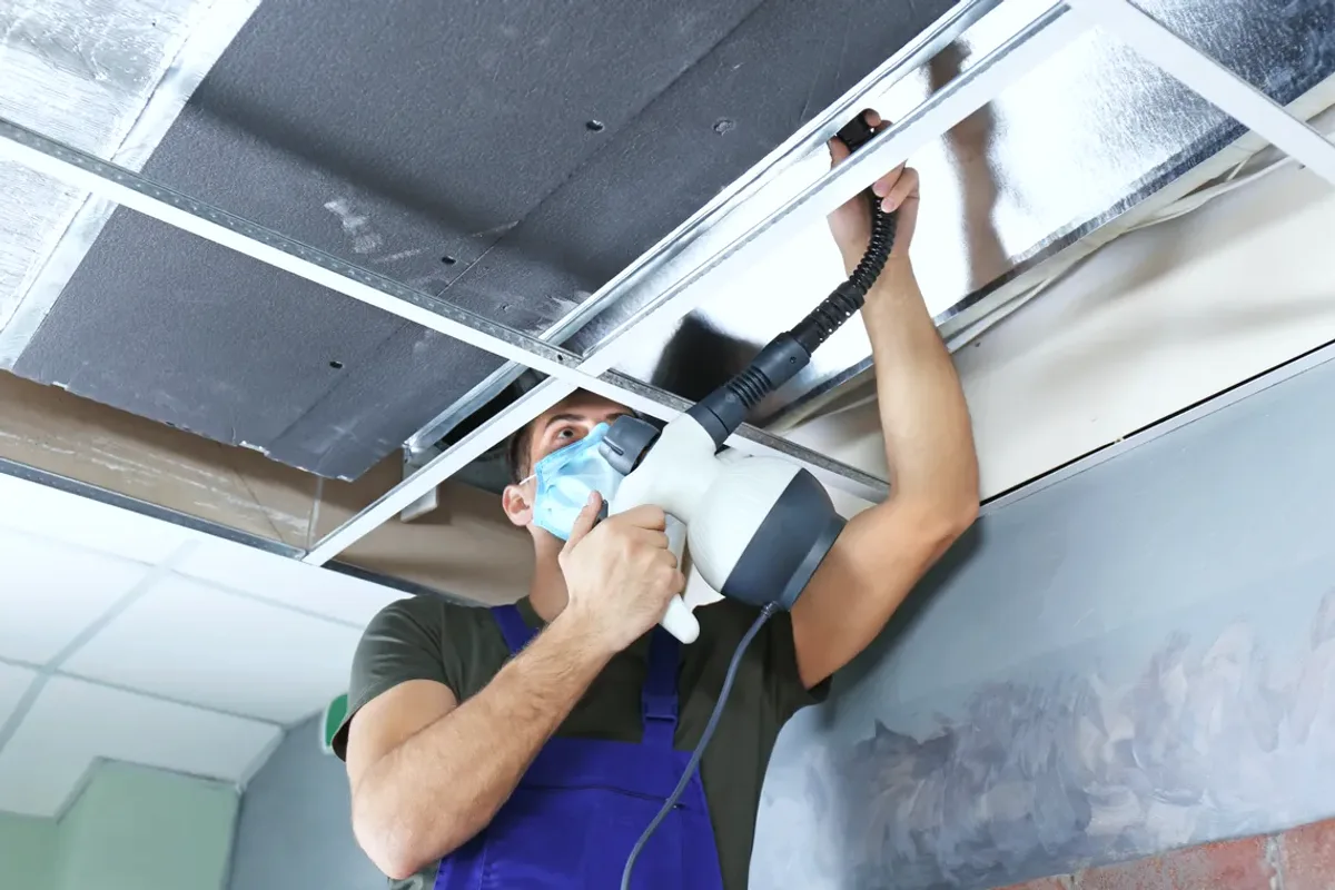 Worker in a mask cleaning a ceiling air duct with a handheld tool