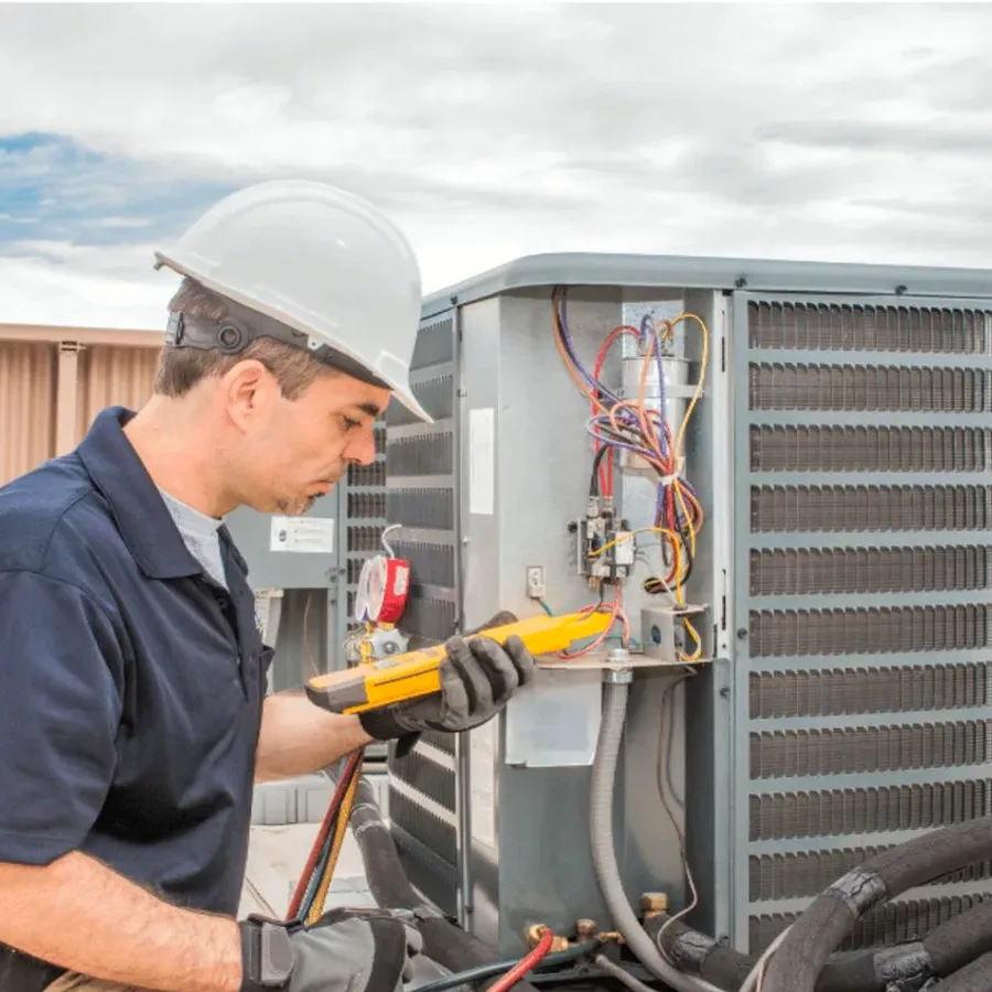 HVAC technician in a hard hat testing an outdoor air conditioner with a multimeter